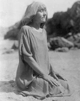 Vintage black-and-white photograph of a woman with short hair and headscarf, kneeling meditatively on a beach