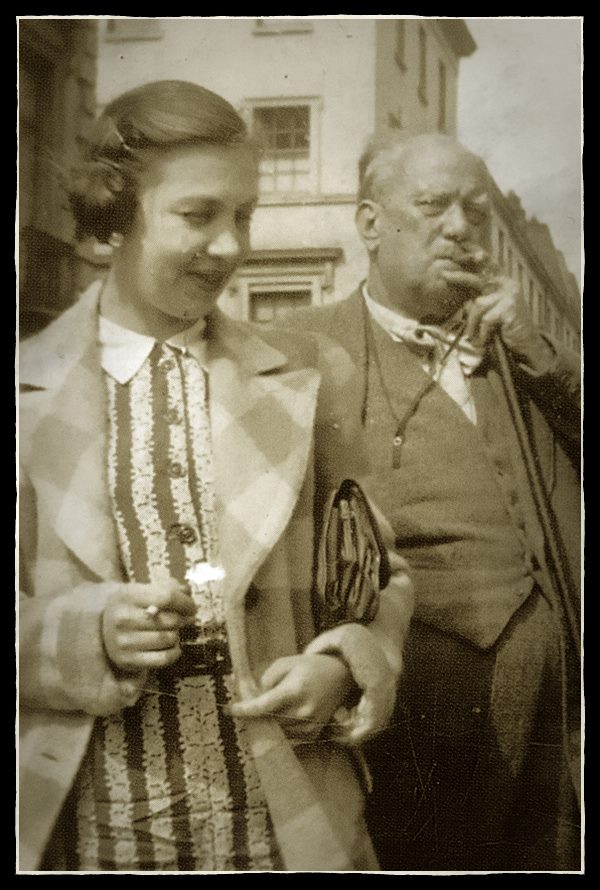 Sepia-toned vintage photograph of a young woman in checkered coat smiling beside older man with pipe