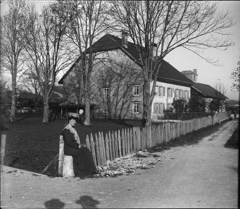 Woman in long dress sitting by fence in front of country house
