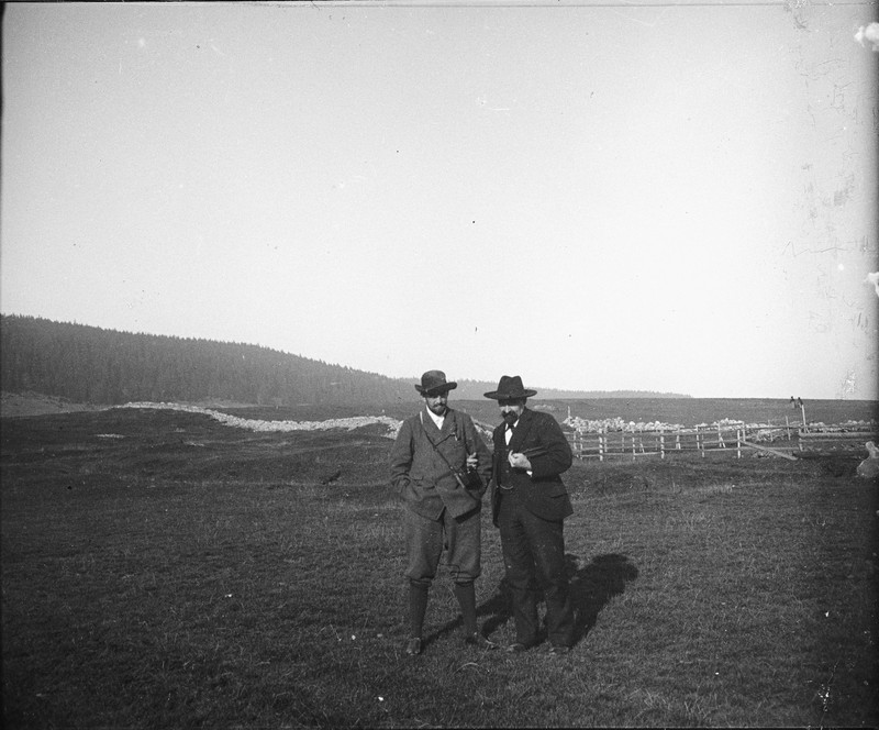 two men in hats and suits standing in a large open field, with a forest and a low stone wall in the distance