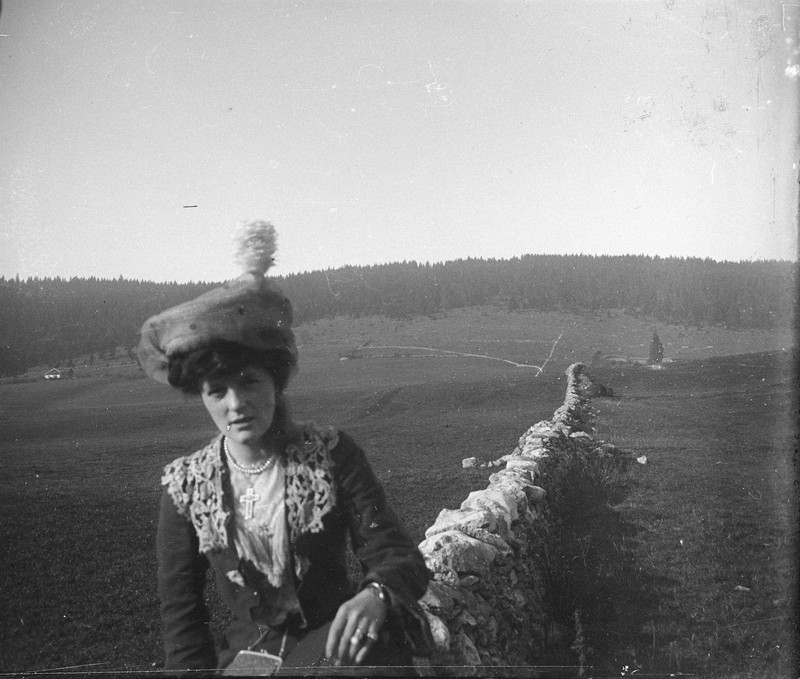 a woman wearing a decorative hat and lace collar, posing outdoors in a field with a low stone wall