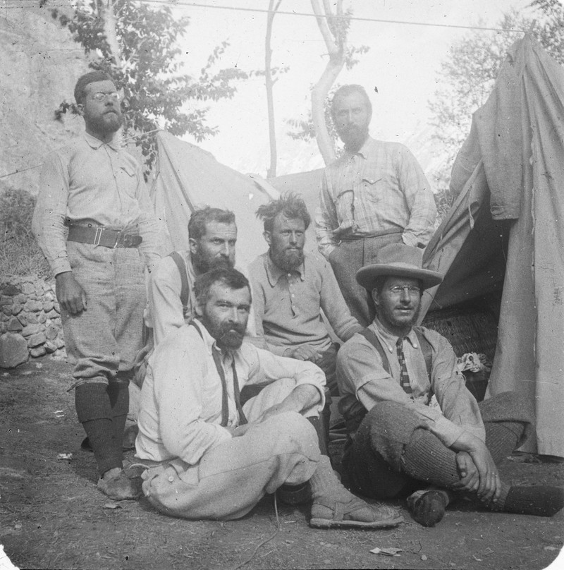six bearded men in early 20th-century outdoor attire, posing together in front of a canvas tent at a campsite