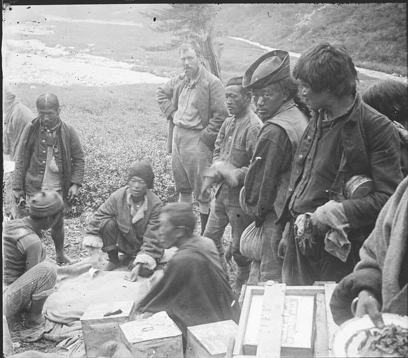 group of men in diverse, rugged attire gathered around several crates in an outdoor, rural setting