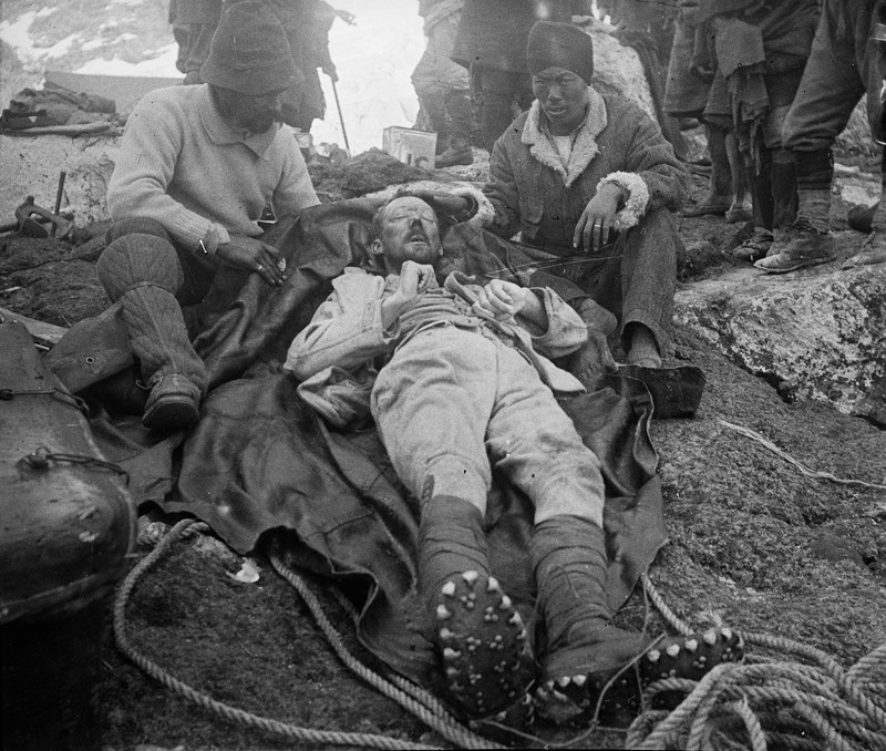 rescue scene on a mountain, showing a man lying on a dark tarp while two others tend to him