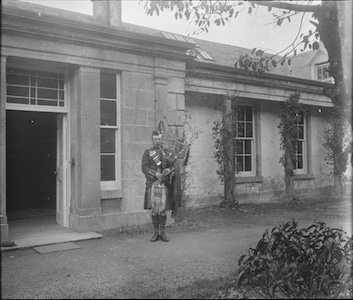 Scottish bagpiper in traditional regalia playing outside a stone building