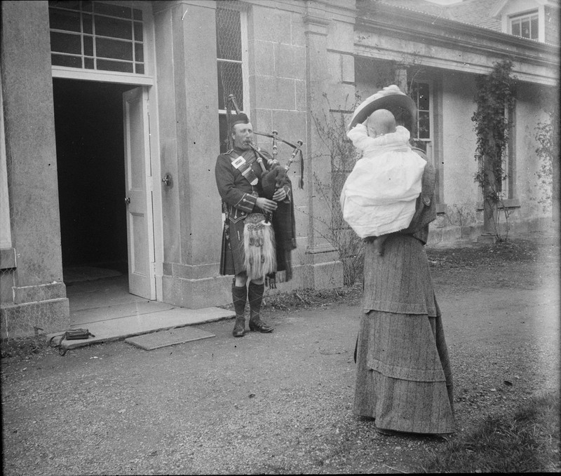 Scottish bagpiper in kilt playing for woman holding baby outside stone building