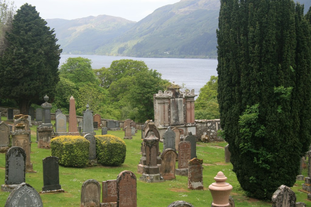 Historic lakeside cemetery with stone monuments, cypress trees, green bushes, misty mountains