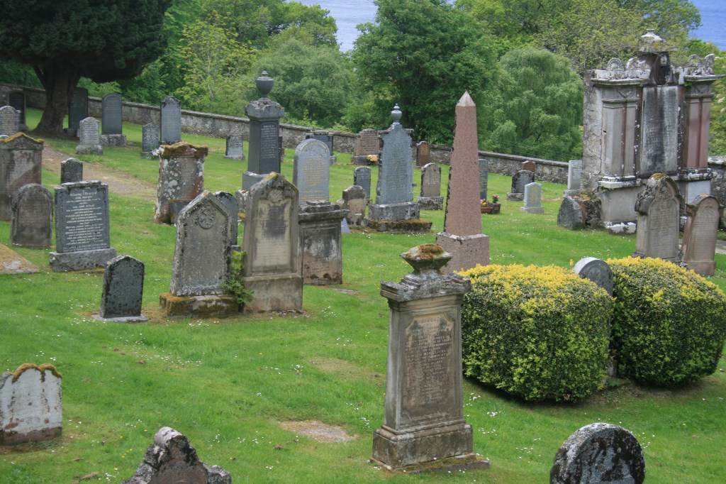 Hillside graveyard with stone headstones, obelisk, monuments, trees, and water view
