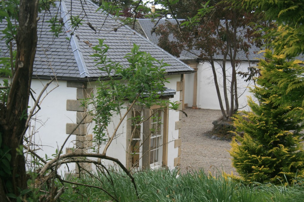 White stone cottage with slate roof, sash windows, quoined corners, surrounded by greenery