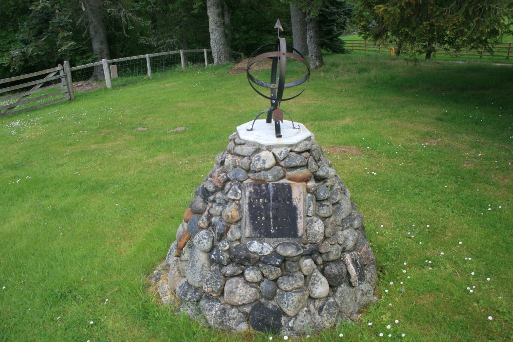 Stone cairn monument in grassy field topped with metal armillary sphere and weathered plaque
