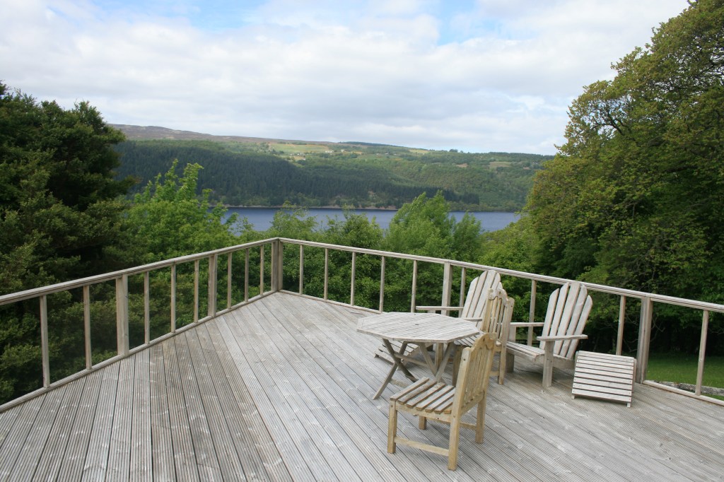 Wooden deck with outdoor furniture overlooking Loch Ness and forested hills under cloudy sky