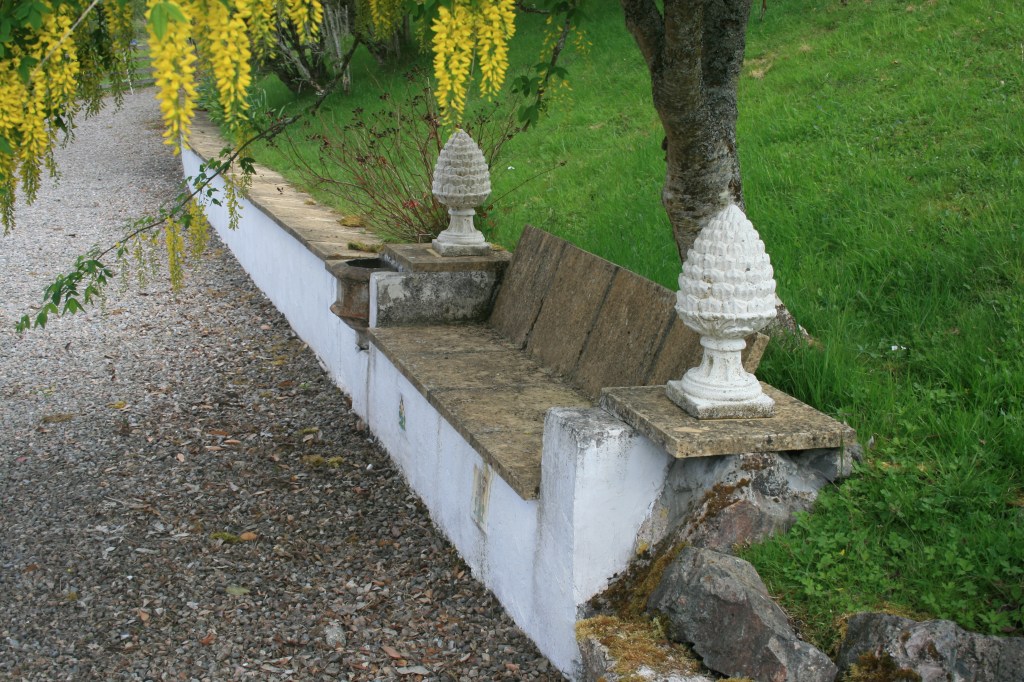 Stone garden bench set into a low white wall with two white pineapple finials beneath a yellow-flowering tree