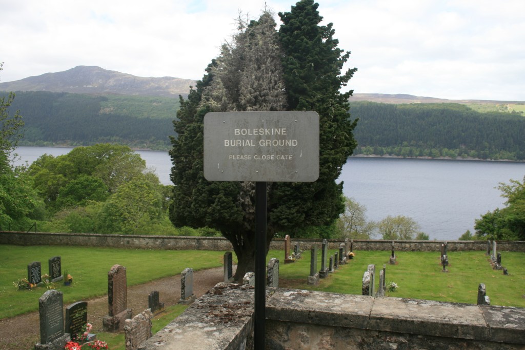 Wide view of Boleskine Burial Ground with metal sign, old gravestones, green lawn, and Loch Ness